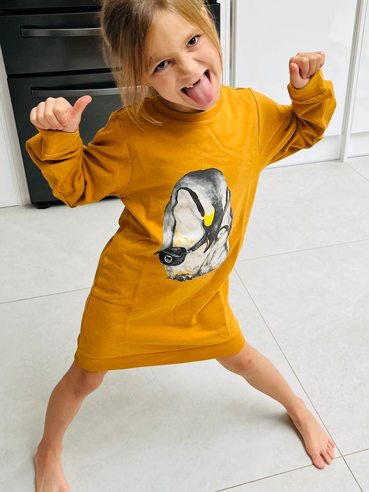 Happy girl giving a thumbs up in The QT mustard Penguin Cuddle organic cotton jumper dress